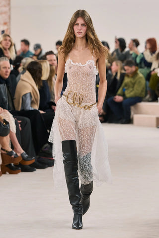 Runway model in a sheer white lace boho festival dress with tall black leather boots.