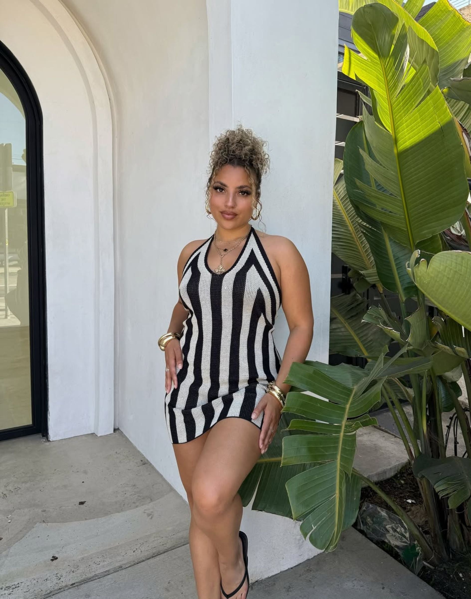 Black and white striped knit halter mini dress styled with gold jewelry by a tropical plant.