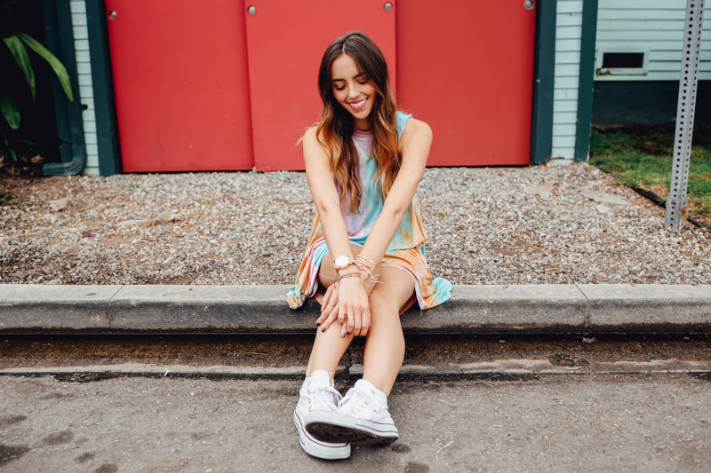 Smiling woman in pastel tie dye tank dress and sneakers seated on a curb.