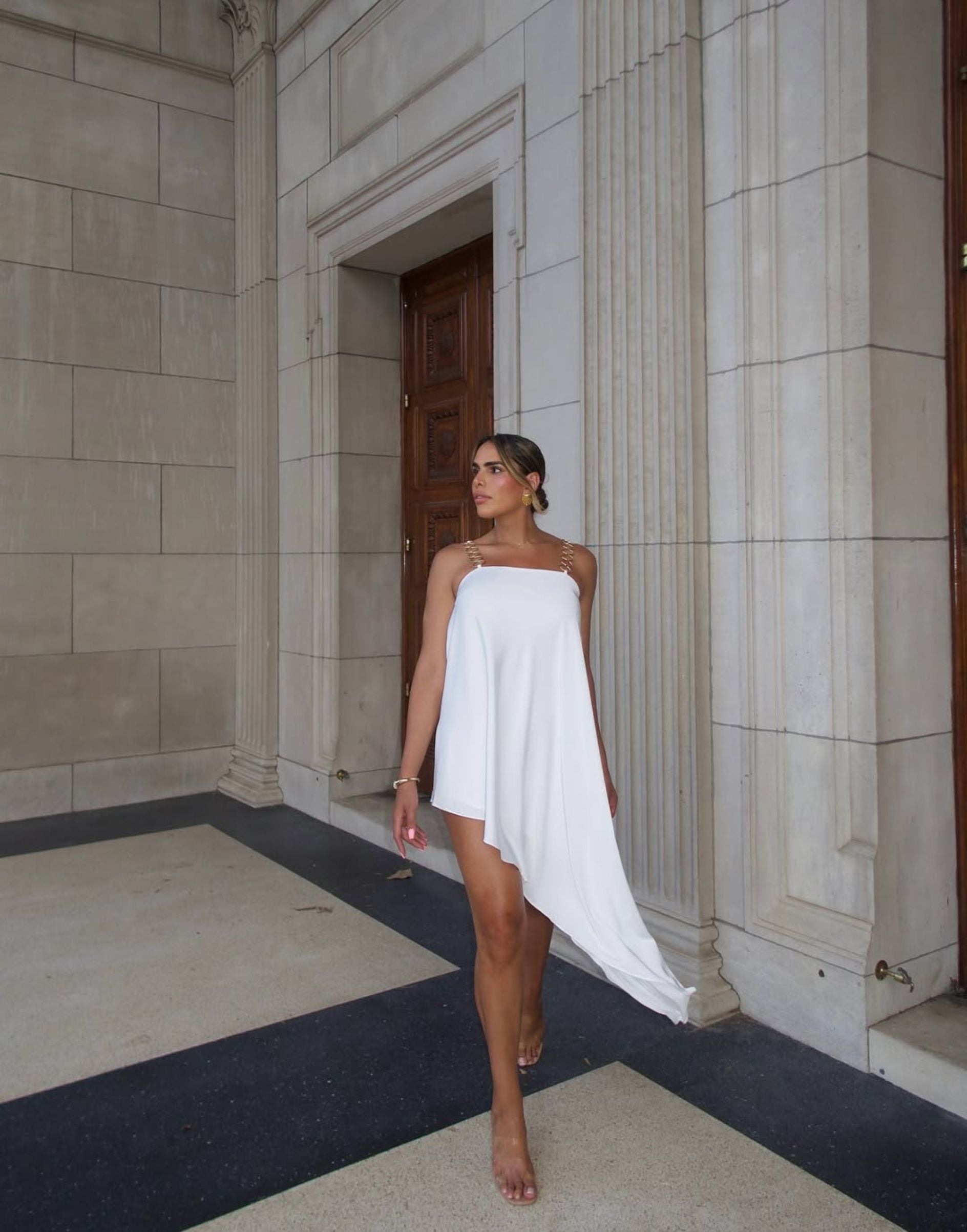 Woman in a white asymmetrical mini dress with gold chain straps beside stone columns.