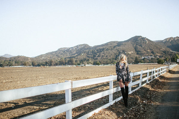 Person in a black floral romper and tall boots leaning on a white ranch fence below SoCal mountains.