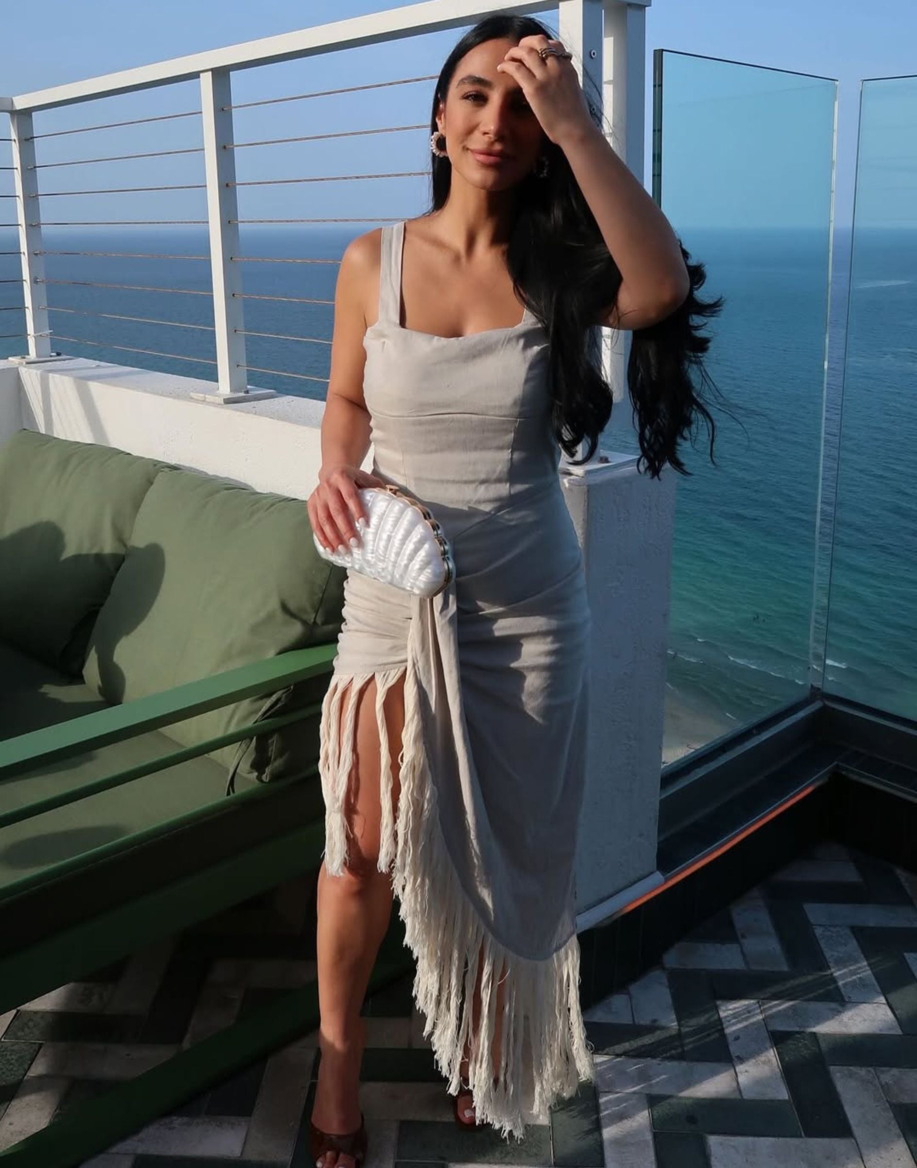 Woman in beige fringe-hem linen resort dress holding a shell clutch on an oceanfront balcony.