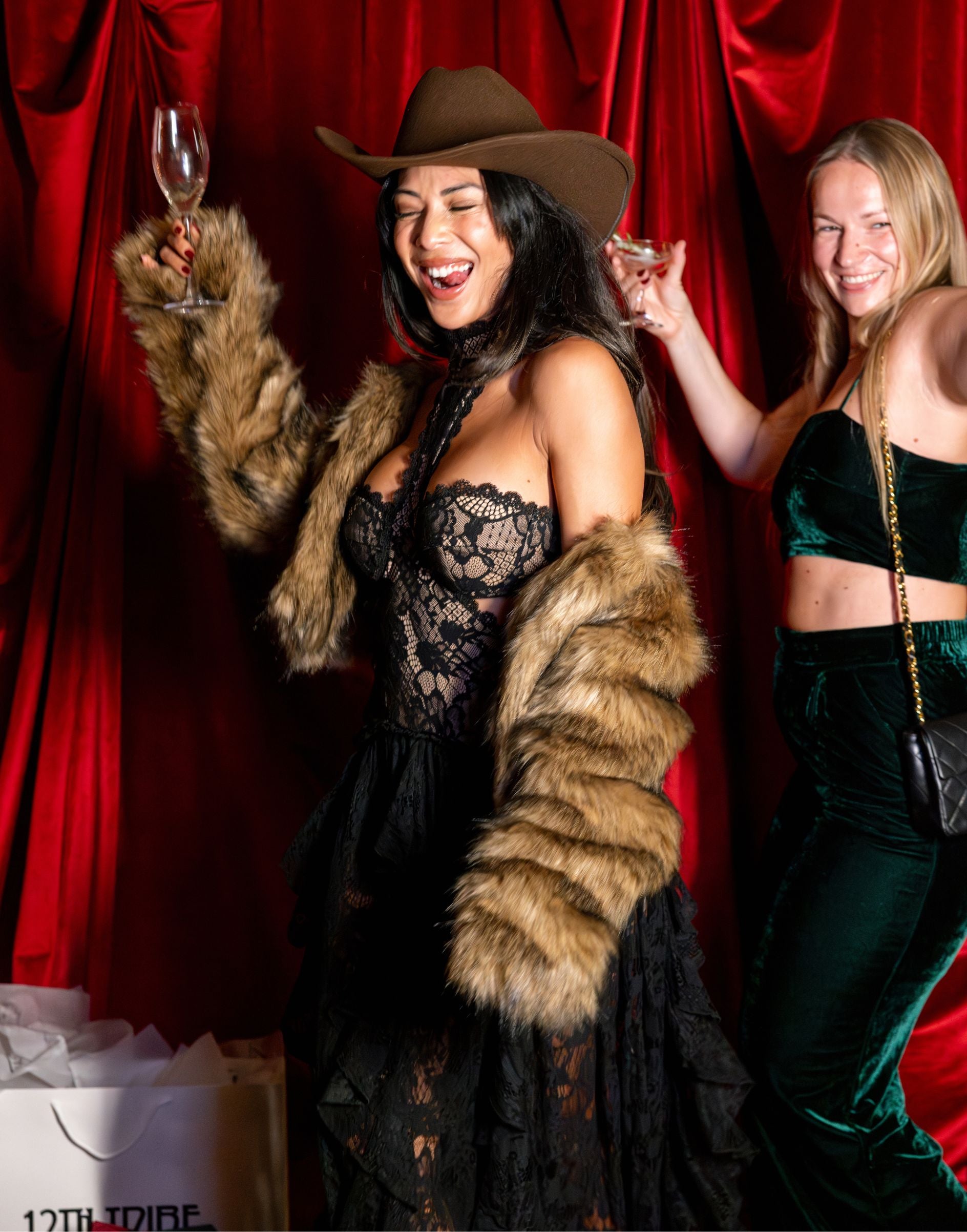 Two laughing guests in black lace and green velvet party outfits raise glasses against red velvet.