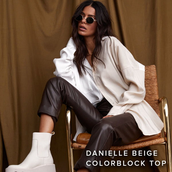 Seated in a woven chair, a woman styles a beige colorblock top with brown leather pants.