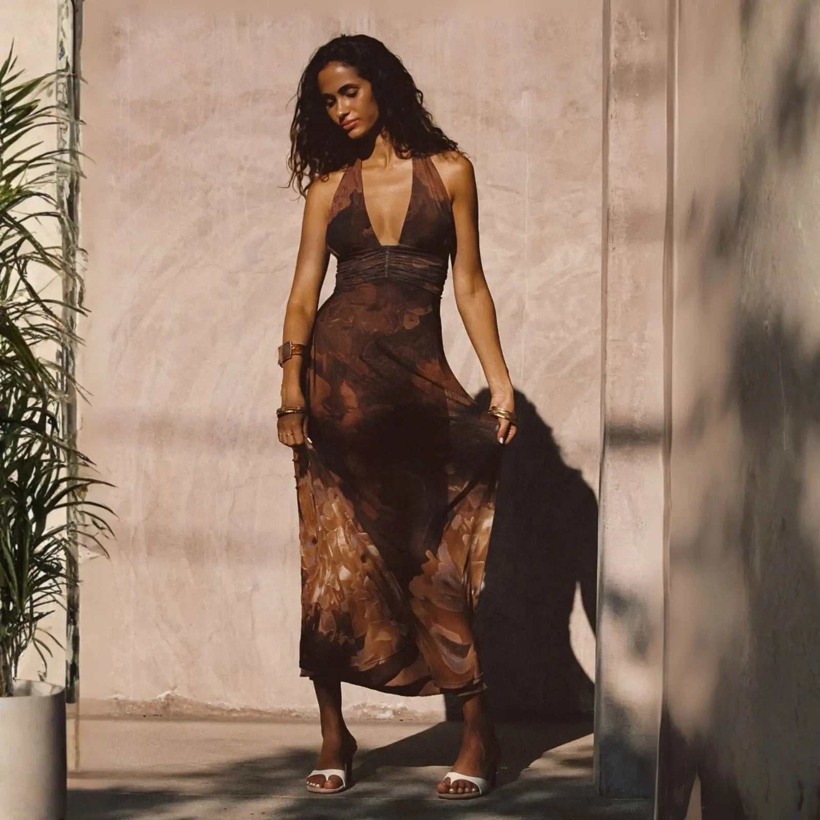 A woman in a sheer brown abstract-print halter maxi dress with gold cuffs stands in dappled sunlight beside a tropical plant, embodying effortless style dresses.