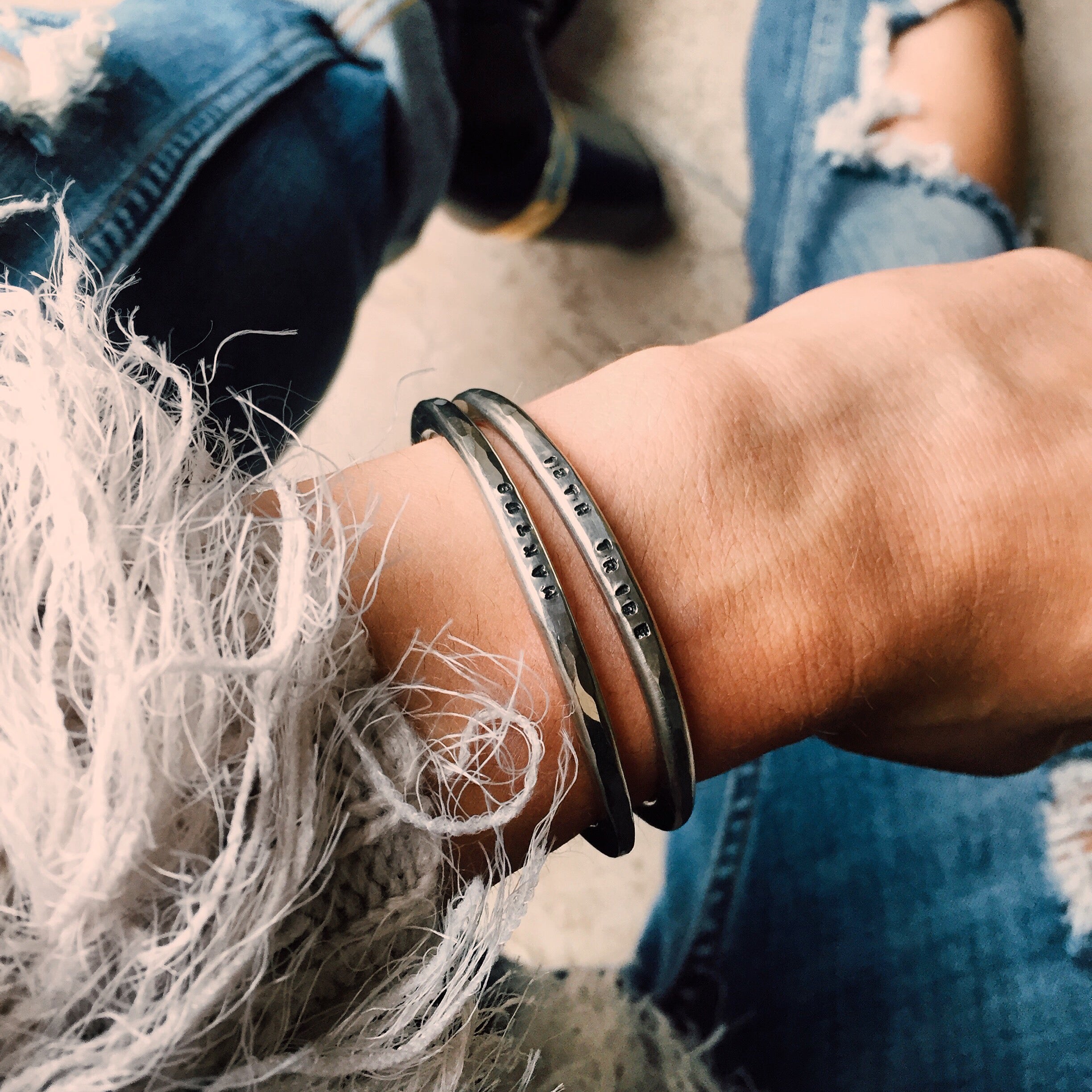 Stacked silver cuff bracelets stamped with text on a wrist beside frayed denim sleeves.