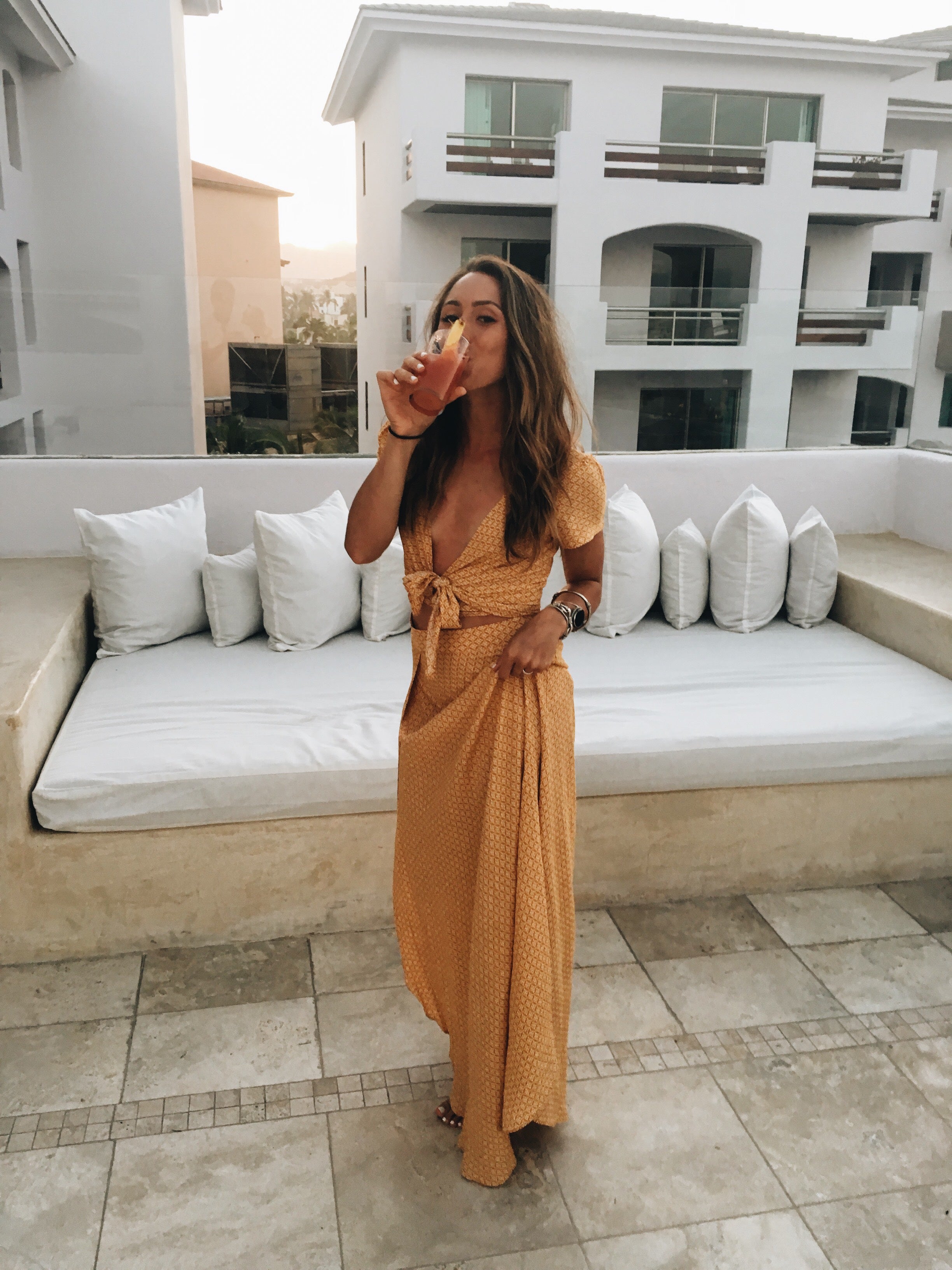 Woman in mustard two-piece resort outfit sips a cocktail on a Cabo San Lucas terrace.