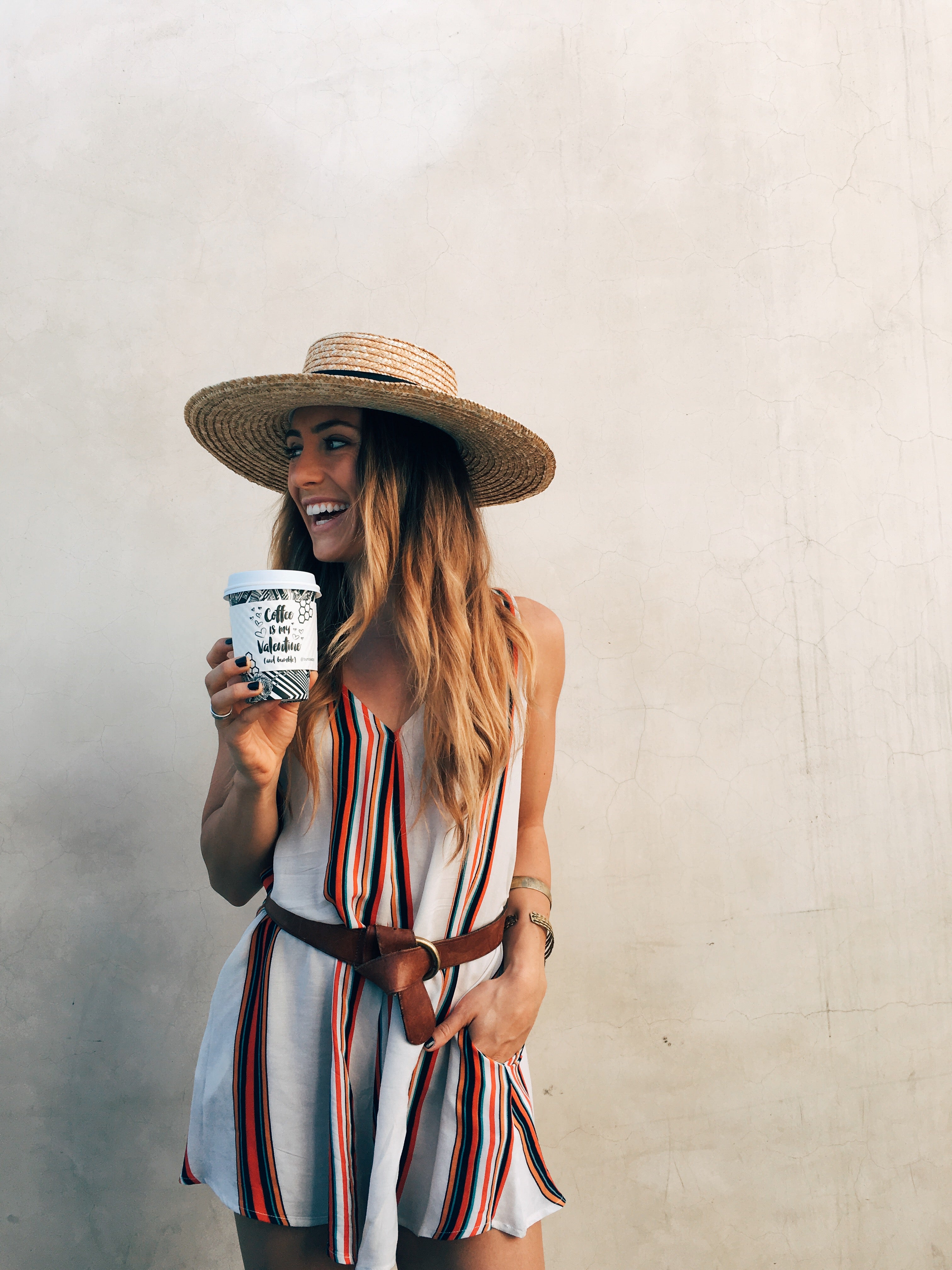 Smiling woman in striped sleeveless romper and straw hat holds a coffee cup outdoors.
