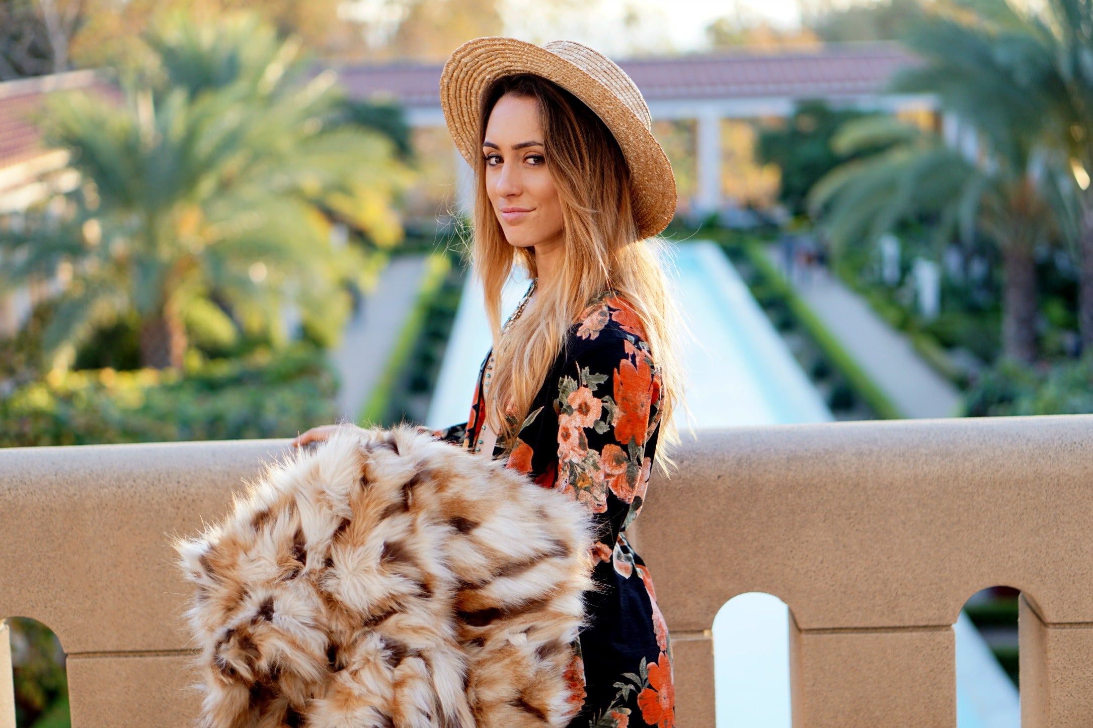 Woman in floral dress and straw hat holding faux fur wrap at a palm-lined resort.