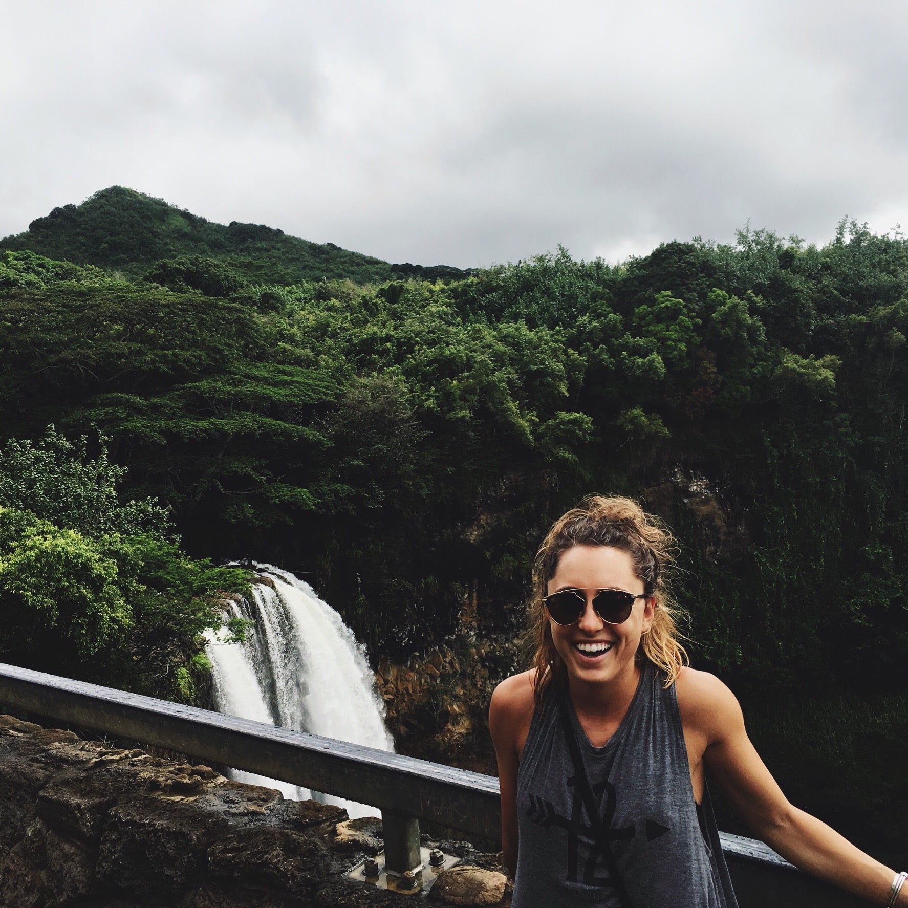 Smiling woman in sunglasses at a Kauai waterfall overlook framed by green mountains.