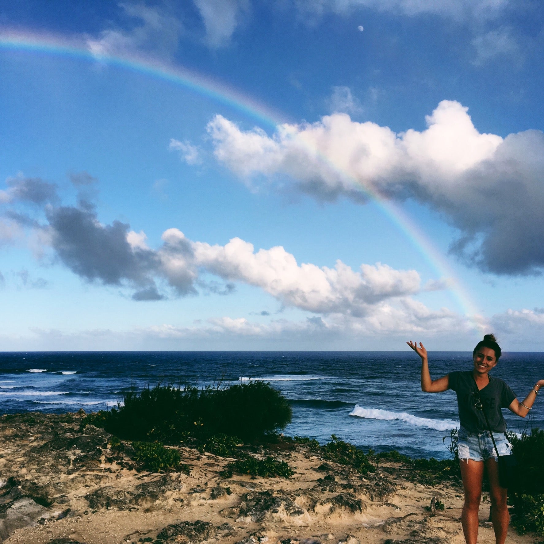 Person waves beneath a rainbow arcing over the rocky Kauai, Hawaii coastline and ocean waves.