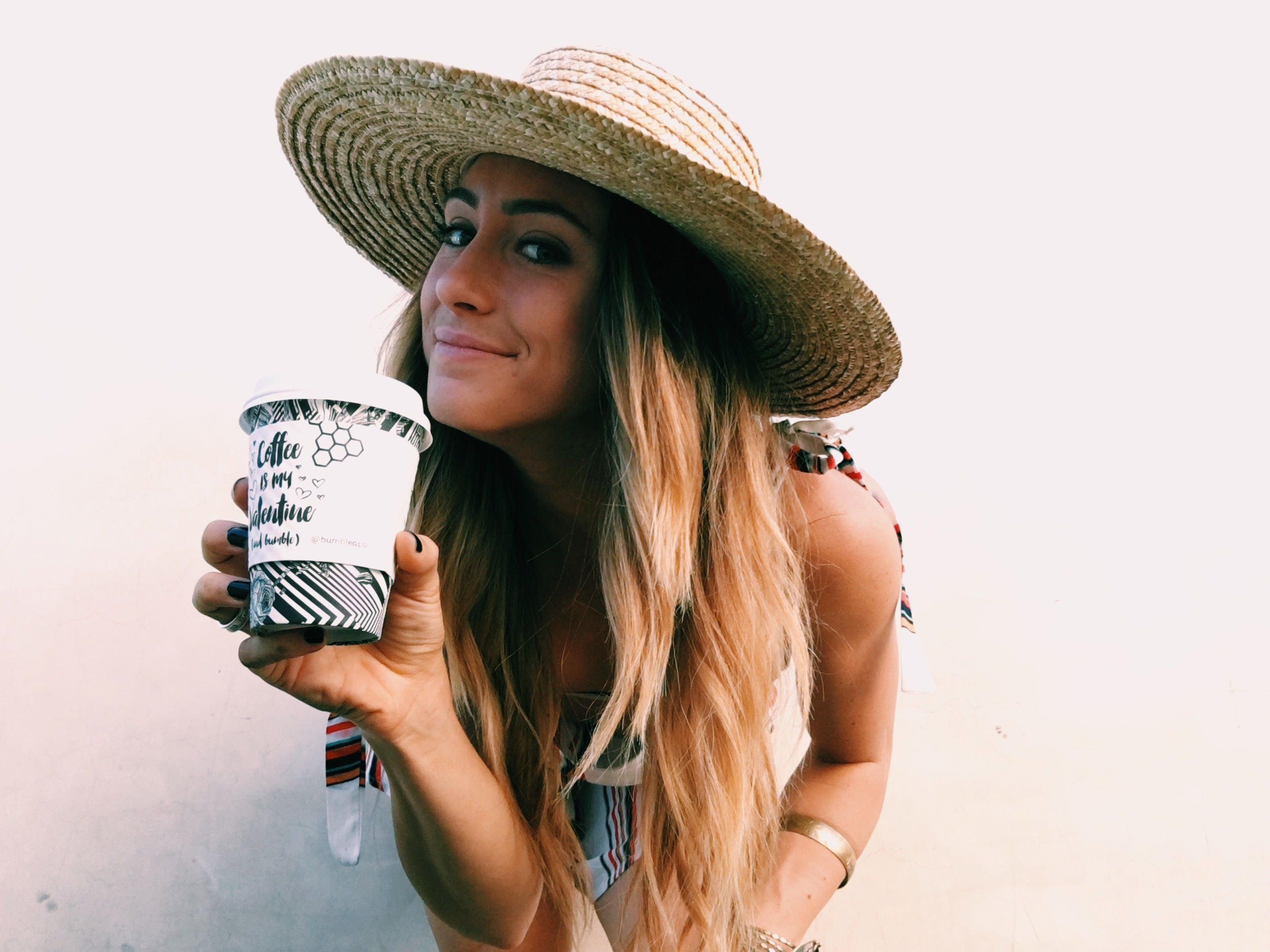 Smiling woman in a straw sun hat holds a printed coffee cup, styled in a summer resort outfit.