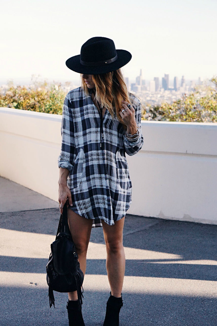 Person in plaid shirtdress and black wide-brim hat with city skyline behind.
