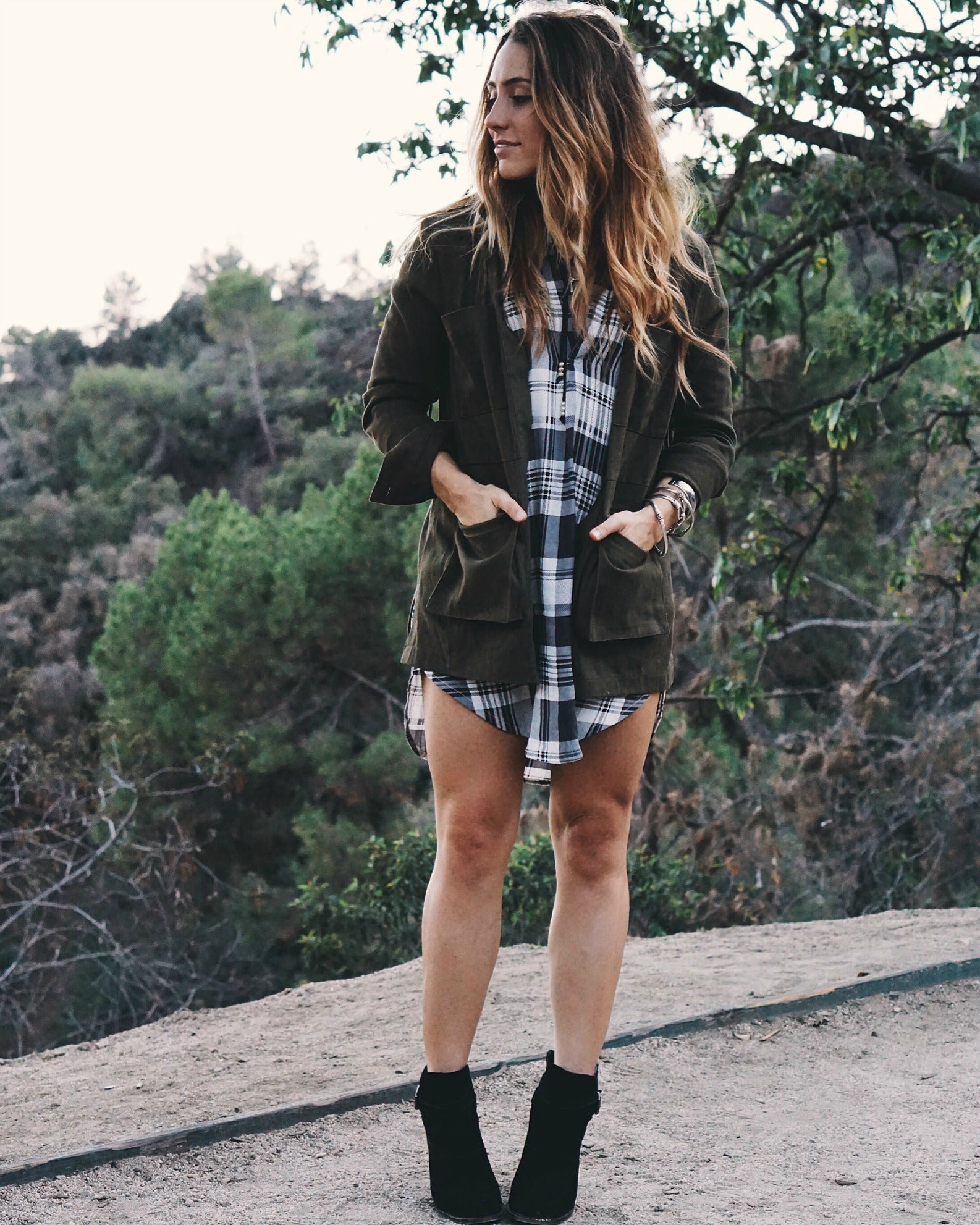 Woman in olive utility jacket over plaid shirt dress with black ankle boots on a trail.