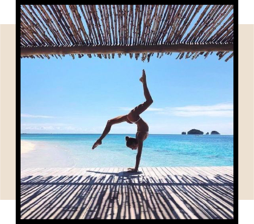Person practicing a handstand yoga pose on a beach deck overlooking turquoise water.