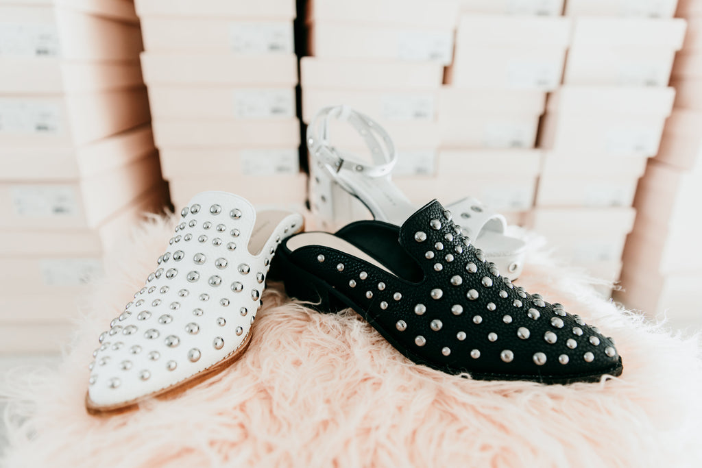 Silver-studded white and black pointed mules resting on pink faux fur beside stacked shoe boxes.
