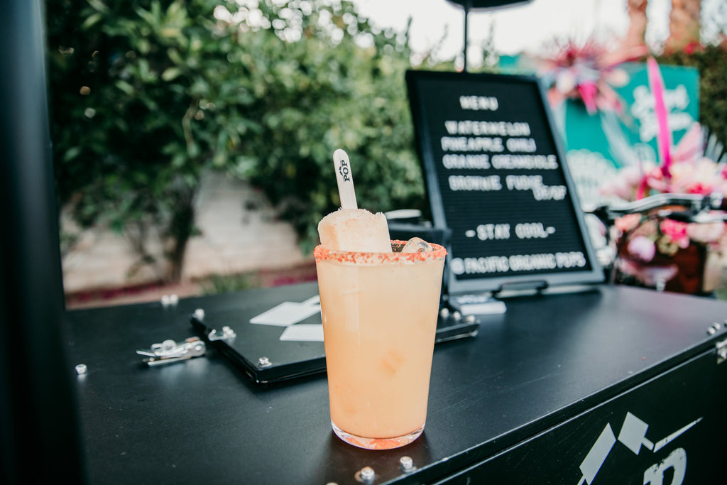 Popsicle-topped cocktail in a salt-rimmed glass beside an outdoor Coachella event drink menu.