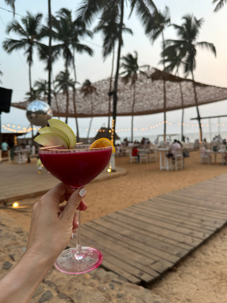 Hand holding a pink cocktail at a palm-lined beach club at dusk in India.
