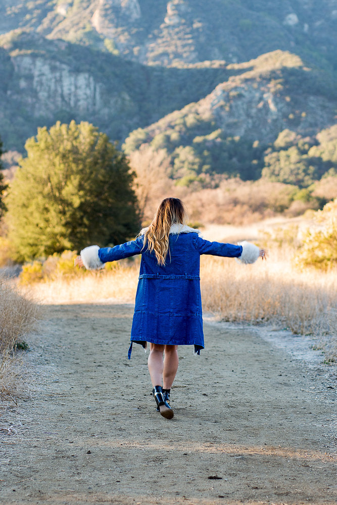 Person with arms outstretched walking a dirt trail toward sunlit mountains in a denim coat.