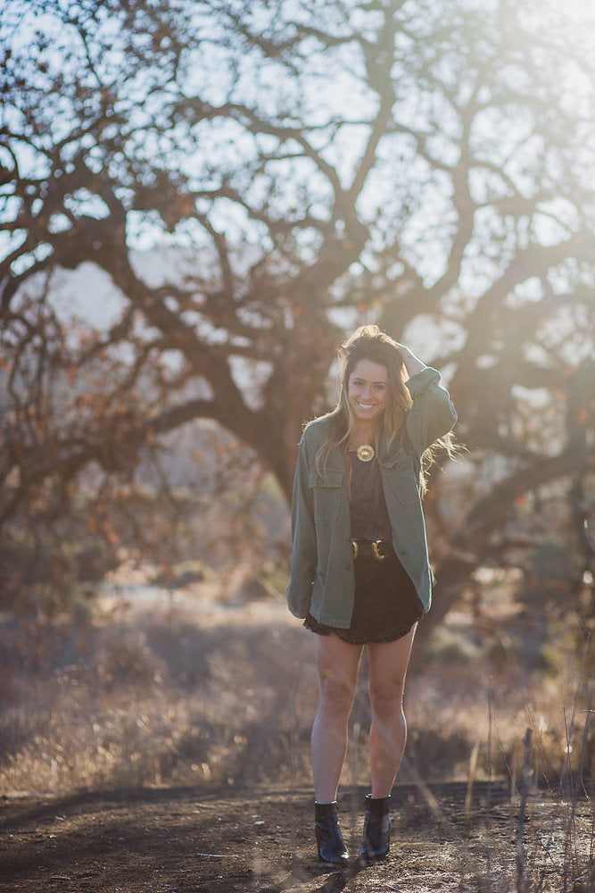 Smiling person in olive jacket and suede skirt, a bohemian festival outfit, in golden sunlight.