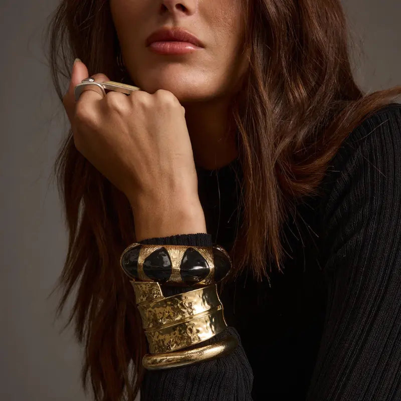 Accessories for a black dress featuring stacked gold cuff bracelets with a hammered texture and a black stone accent worn over a ribbed black knit top with a silver statement ring