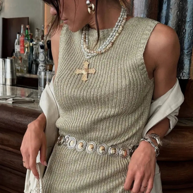 Accessories for dresses layered with pearl and shell multi-strand necklaces, a gold cross pendant, chunky gold hoop earrings, a concho belt, and silver cuff bracelets over a metallic gold ribbed knit dress at a bar