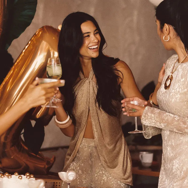 Bachelorette clothes in gold shimmer including a halter crop top and matching pants worn while laughing with friends holding cocktails at a balloon-filled celebration