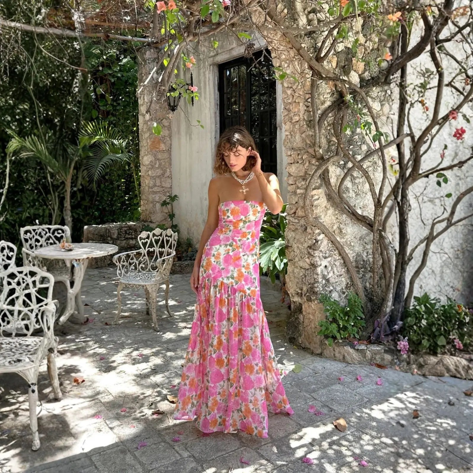 A woman in a strapless pink and orange floral maxi dress with a layered necklace poses in a sun-dappled stone courtyard, wearing one of the most chic vacation dresses.