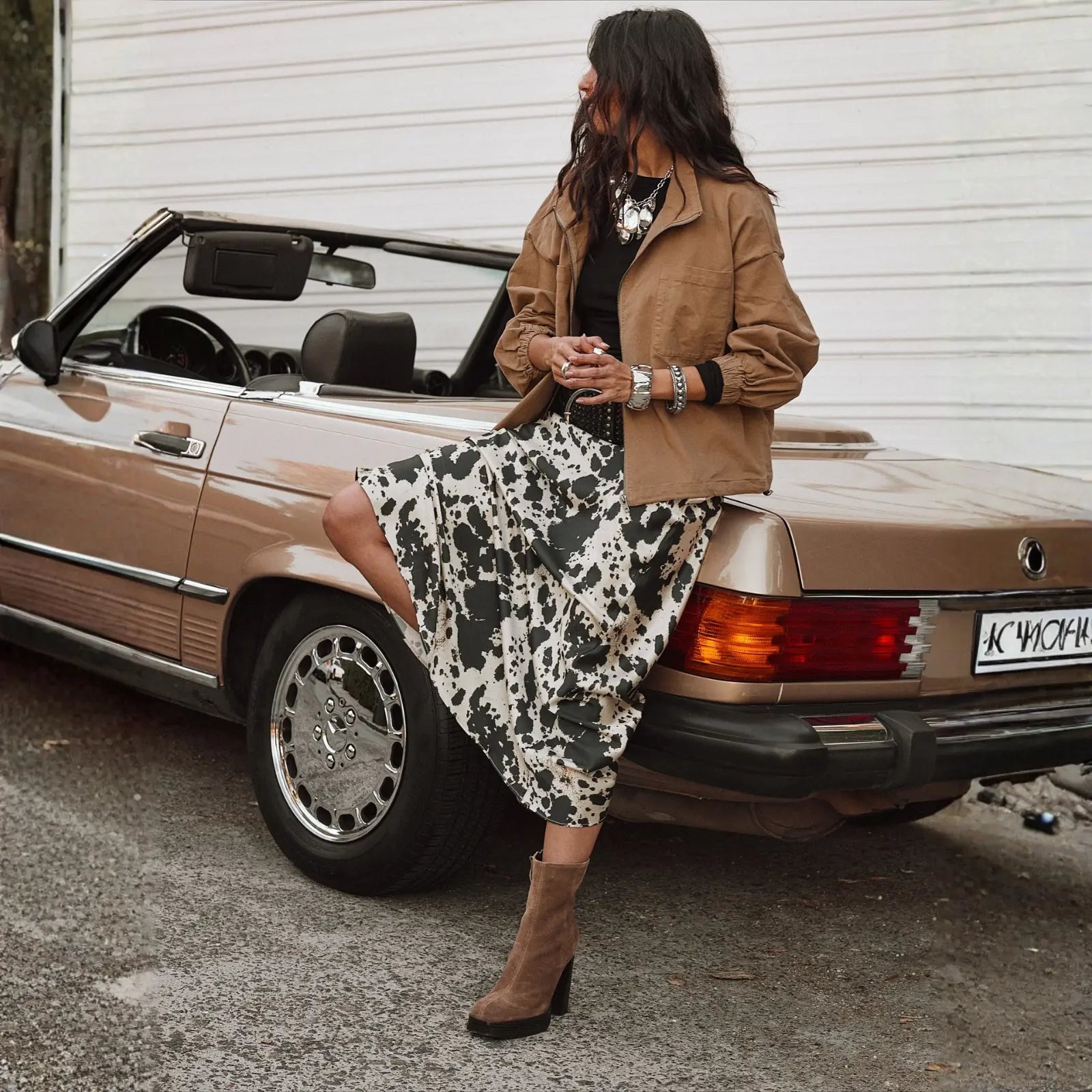 A woman in a cow-print midi skirt, tan jacket, and block-heeled boots leans against a vintage convertible, wearing a chic vacation dress-and-jacket ensemble.