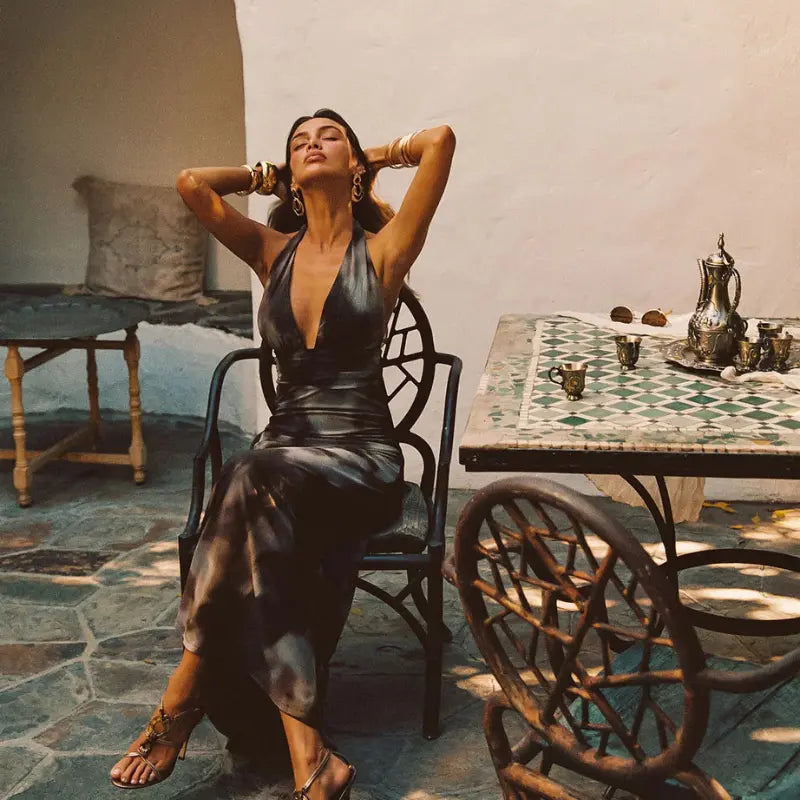 Long sheer maxi dress in metallic charcoal with a deep halter neckline and flowing silhouette styled with gold cuff bracelets at a Moroccan mosaic tile table
