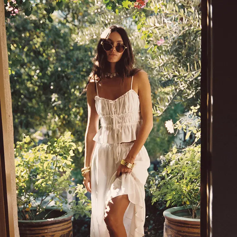 Luxury resort wear in an all-white ruched cami top and flowing ruffle maxi skirt with gold bangles and sunglasses in a sun-dappled garden setting