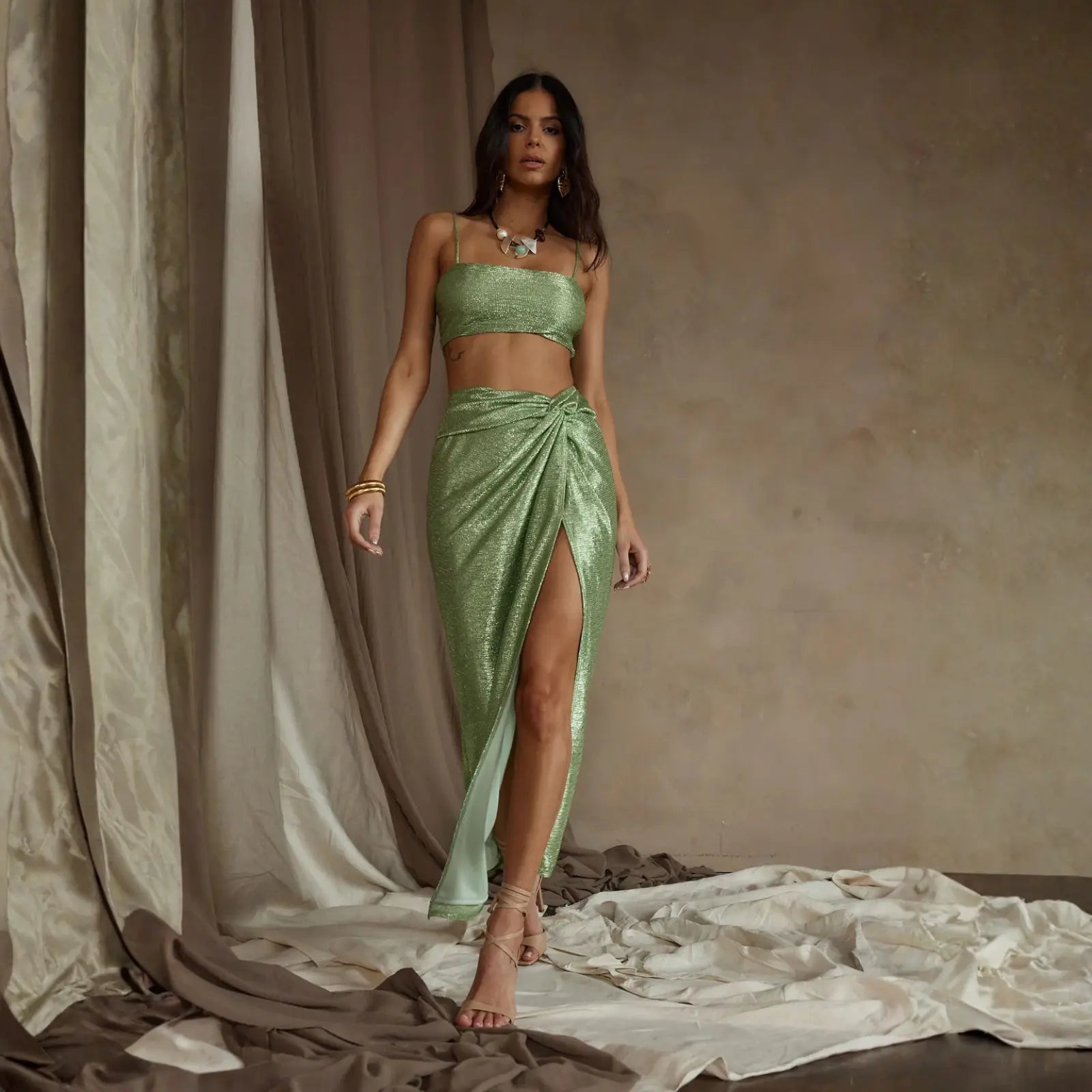 A woman in a sage green metallic crop top and knotted wrap maxi skirt with gold accessories, a glamorous take on luxury summer wear.