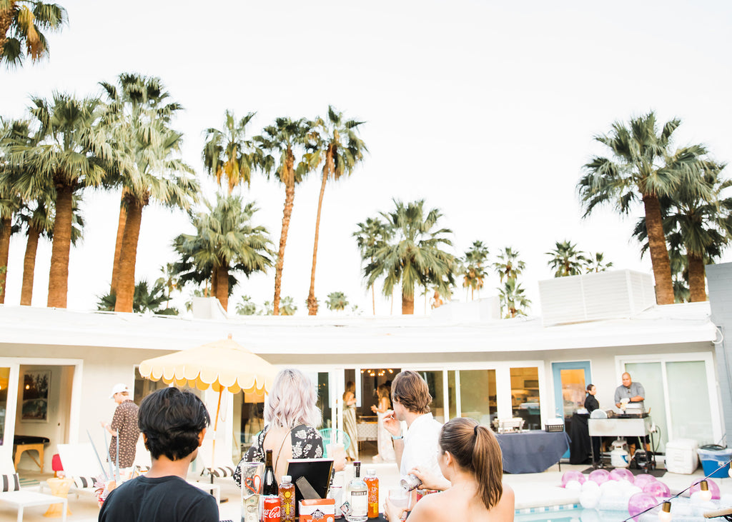 Guests mingling at a Coachella poolside party with palm trees and a yellow umbrella.