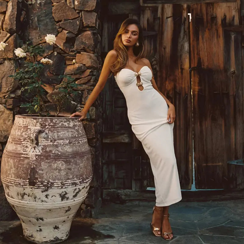 Resort dresses for women in a white strapless maxi with gold ring cutout details and a body-skimming fit styled with gold heels beside a rustic stone wall and wooden door