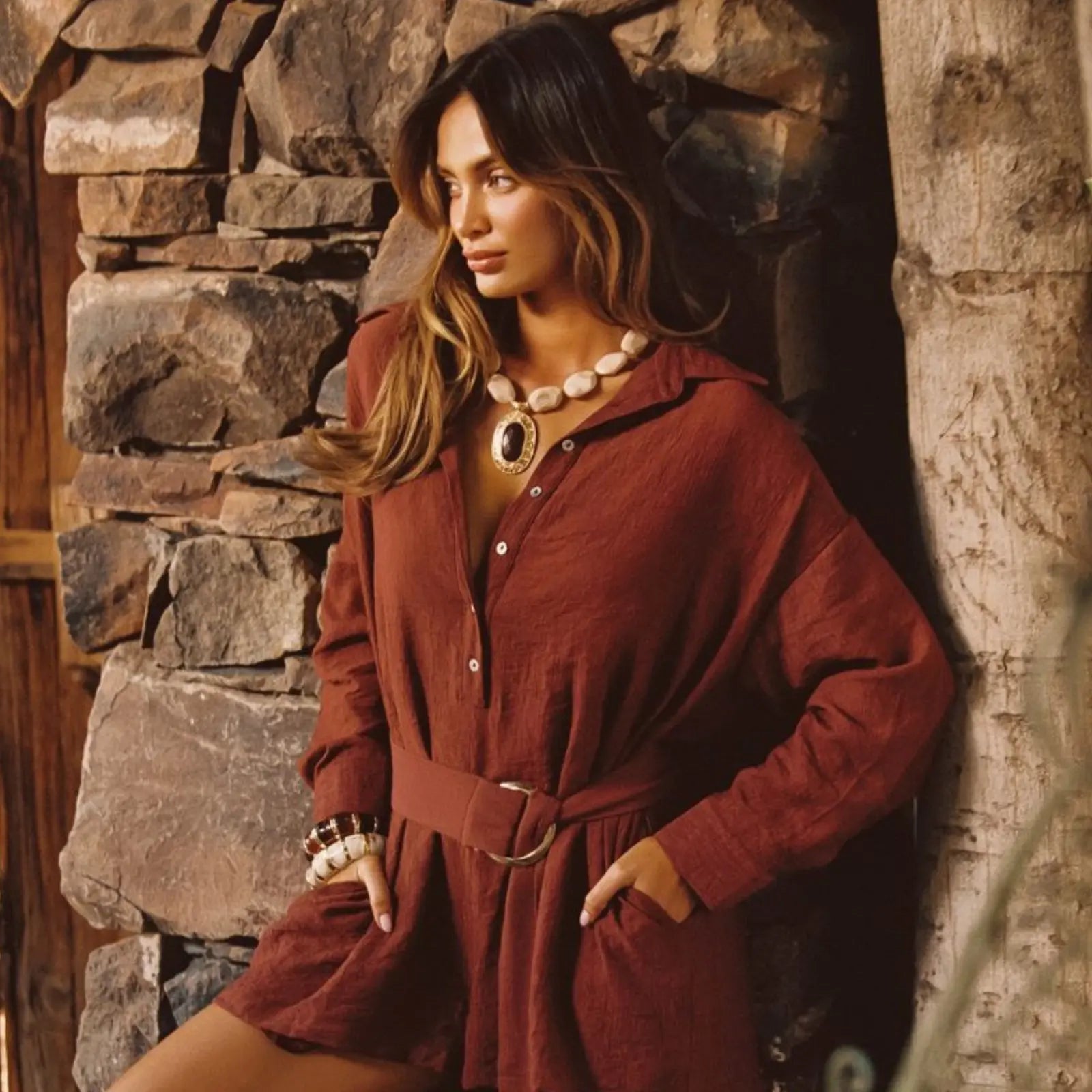 A rust-colored belted linen shirt dress layered with statement jewelry against a rustic stone wall, a relaxed yet polished resort outfit for women.