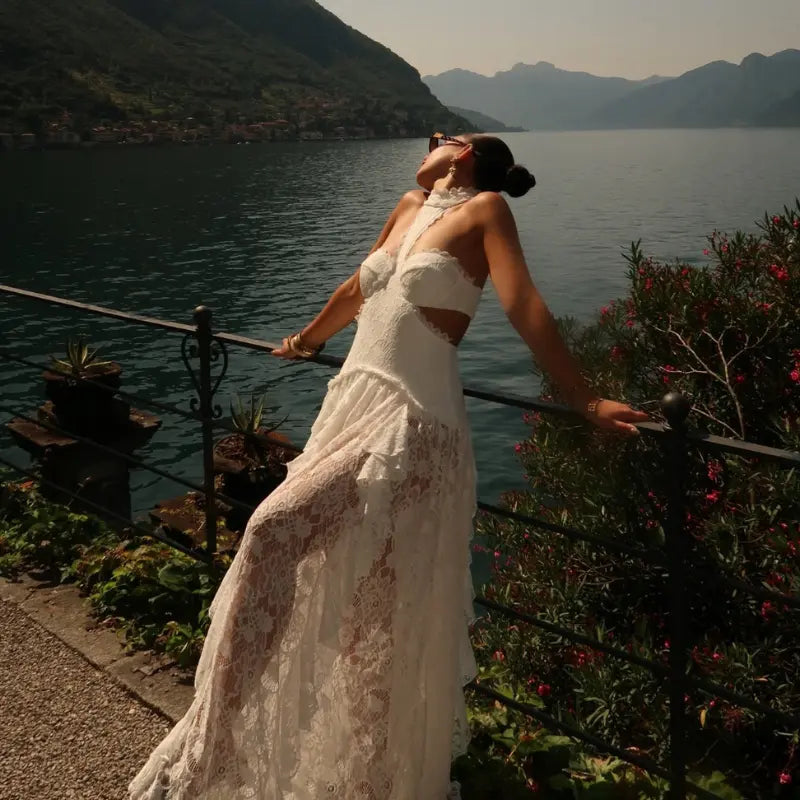 Stylish vacation outfit featuring a white lace halter cutout maxi dress worn lakeside with mountains in the background on a sunny European getaway