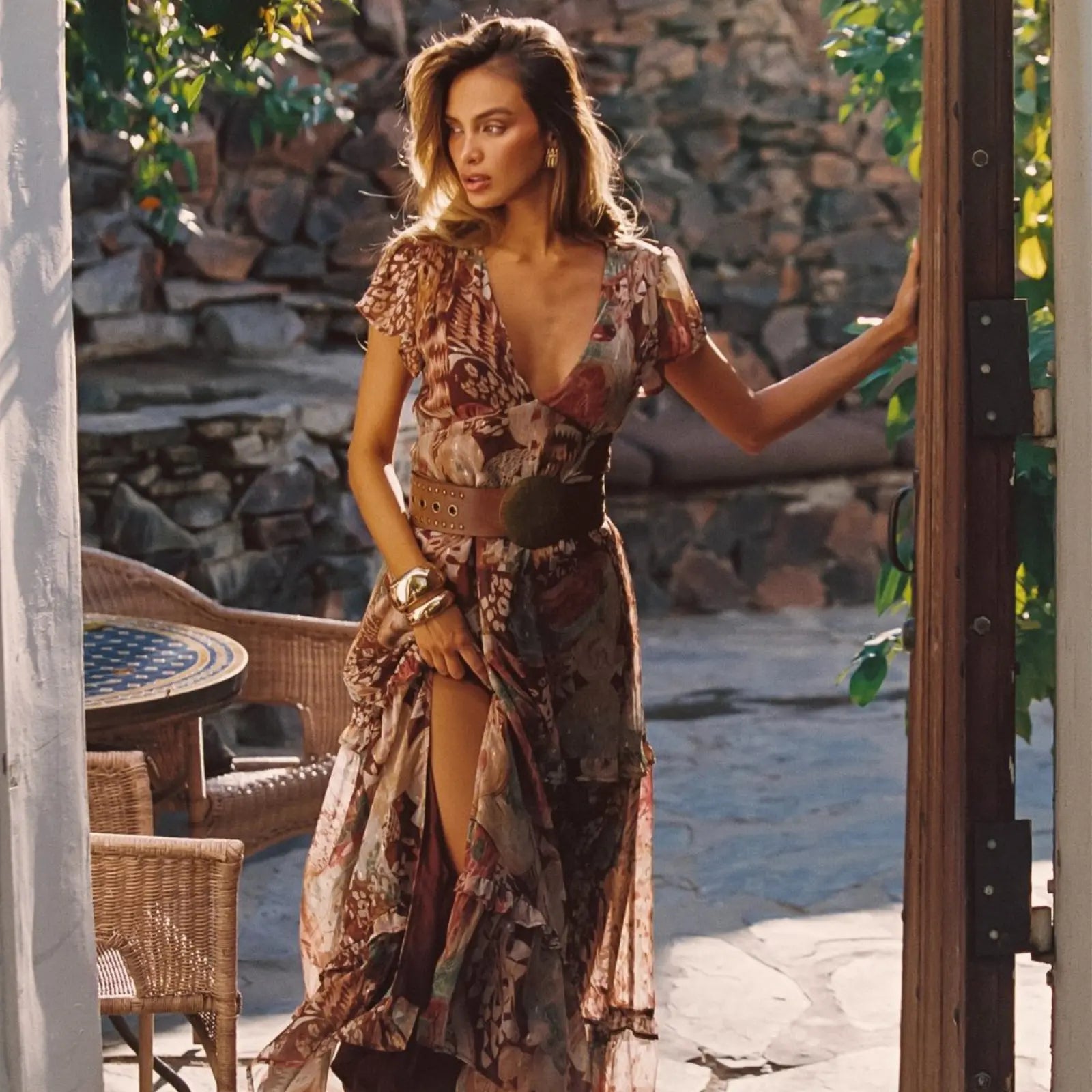 A woman in a belted boho floral maxi dress with flutter sleeves stands in a sun-drenched stone courtyard, showcasing effortlessly stylish vacation outfits.