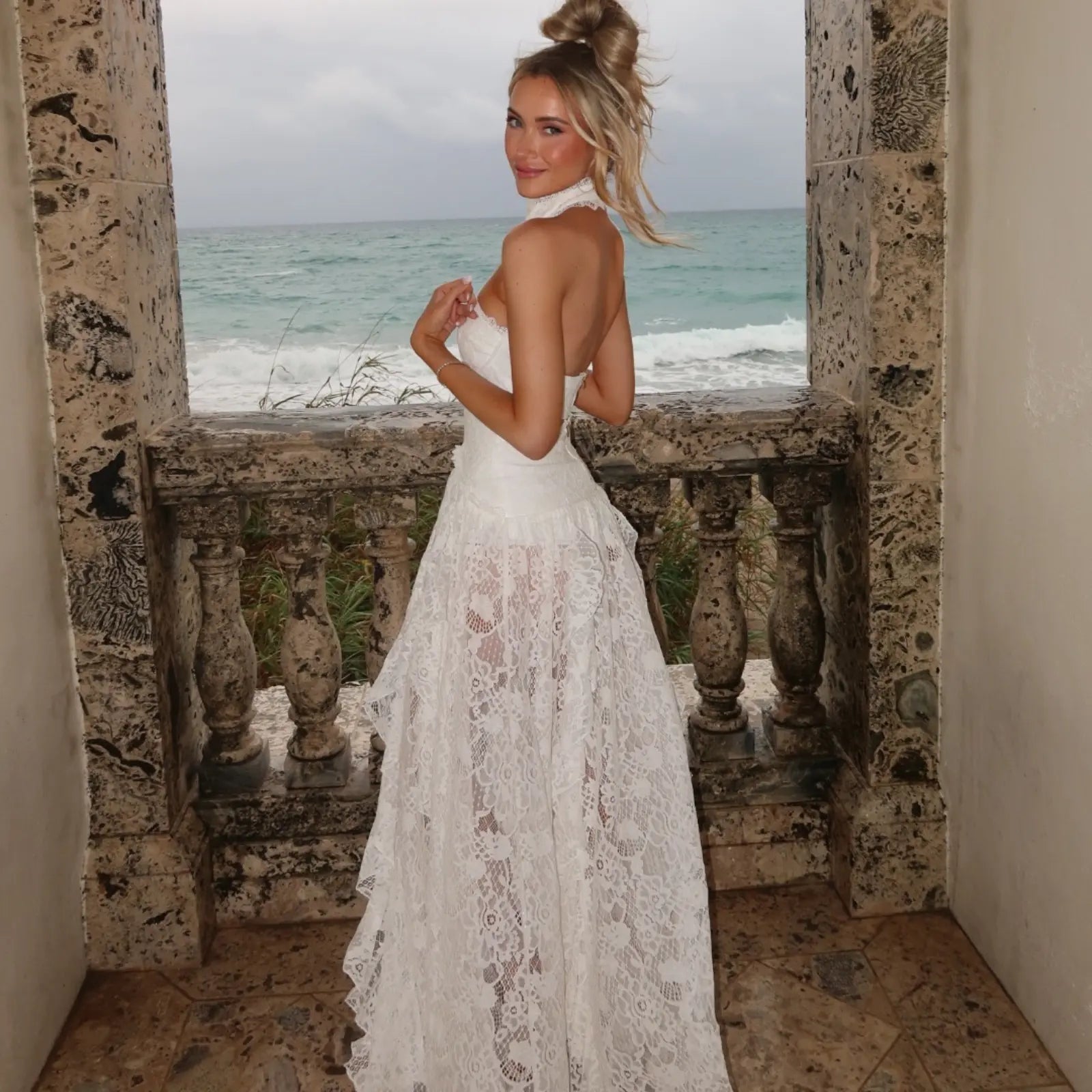 A woman in a backless white lace halter gown poses on a stone ocean-view balcony, wearing a stunning piece of summer vacation clothing.