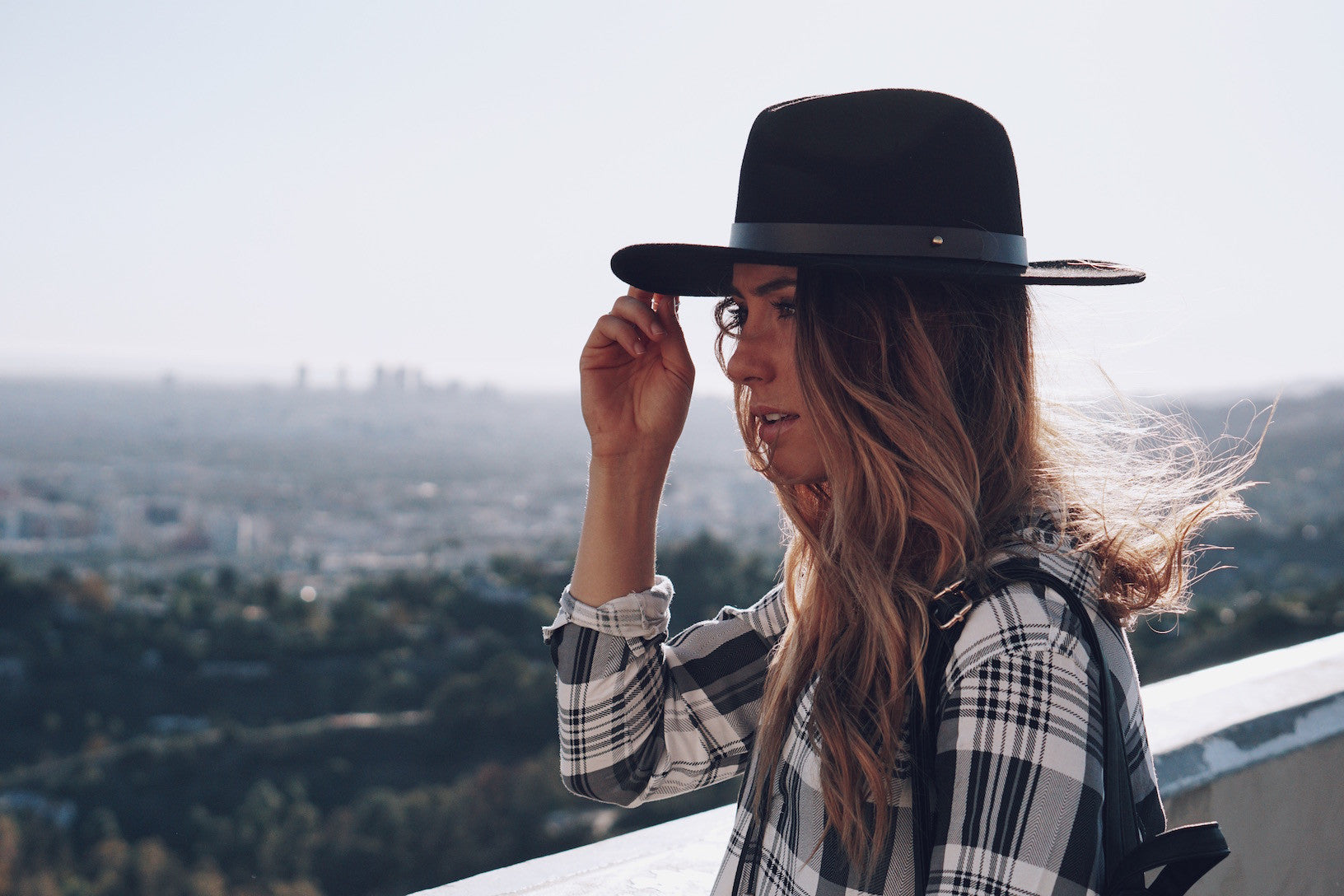 Person in black felt hat and plaid shirt overlooking a hazy city skyline.