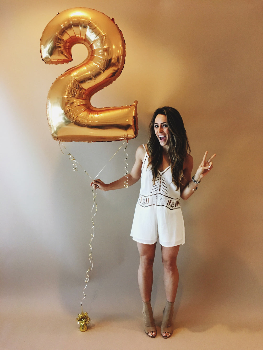 Smiling woman in a white cutout romper holds a giant gold number two balloon.