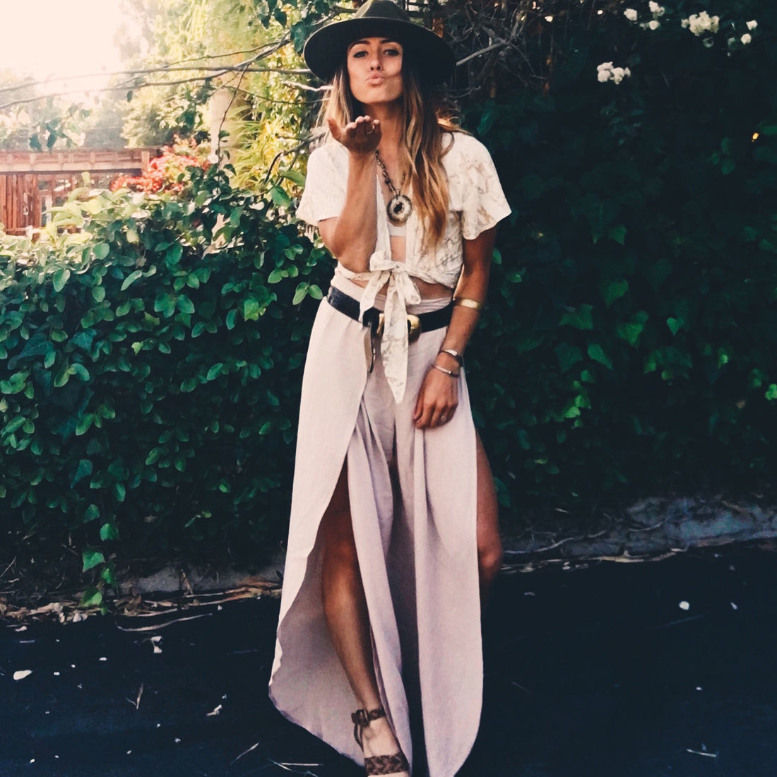 Person in boho tied lace top, maxi skirt, and felt hat blowing a kiss outdoors.